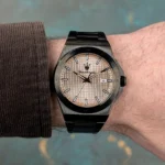 Black metal wristwatch with a beige textured dial, black hands, and a date window showing 8 on the wearer's left wrist.