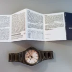Metal wristwatch with a pale dial resting on a gray surface beneath an open instruction manual.