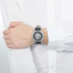 Wristwatch with a visible mechanical movement on a black leather strap worn by a person in a white shirt surface view of the wrist.