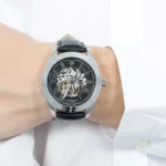 Skeleton wristwatch with visible gears on a black dial, silver case, and black leather strap worn on the wrist. Maserati branding visible at 6 o'clock edge.
