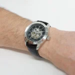 Skeleton wristwatch with visible gears, silver case, black dial with Roman numerals, and black leather strap on a wrist.