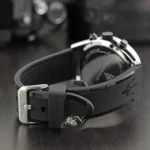 Close-up of a black rubber watch strap with a metal buckle on a wristwatch, showing the back case and crown edge in the background.