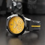 Wristwatch with a vibrant amber dial, black bezel, and a black strap with a yellow stripe detail on a dark surface.