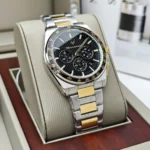 Two-tone stainless steel chronograph wristwatch with a black textured dial and gold-accent tachymeter bezel, resting in a beige display case.