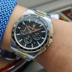 Two-tone stainless steel chronograph wristwatch with a black textured dial and gold accents on the wearer's wrist.