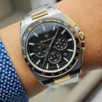 Two-tone wristwatch with a black textured dial, gold indices, and three subdials on a silver-gold metal band. Tachymeter bezel visible around the face.