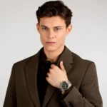 Portrait of a man in a brown blazer and black shirt, adjusting his lapel while wearing a silver and black wristwatch.