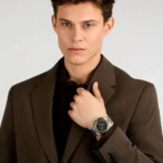 Man in a brown suit adjusts a silver metal wristwatch on his left wrist, posing against a neutral background.